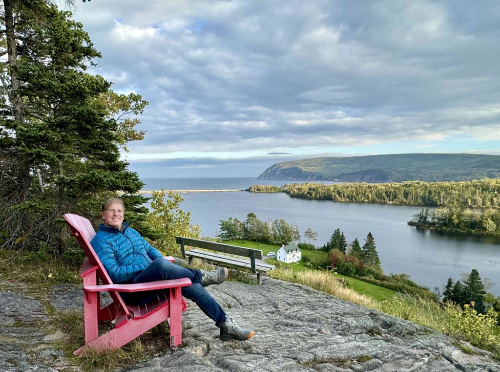 man sitting on red adirondack chair overlooking costal view of the cabot trail