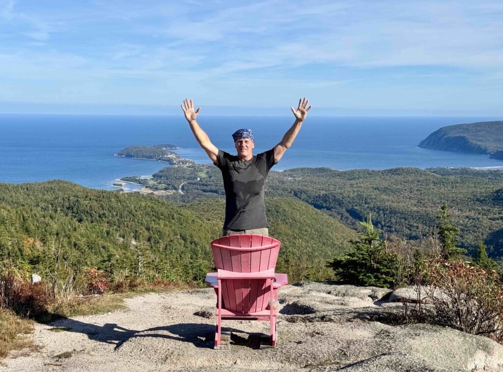 man standing on rocks with hands in the air with a coastal view of the Cabot Trail in the background