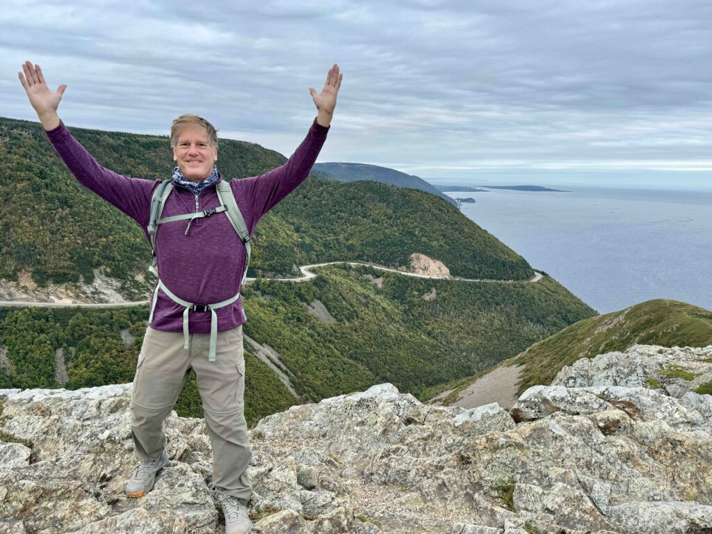 man standing on rocks with arms in the air with the cabot trail in the background